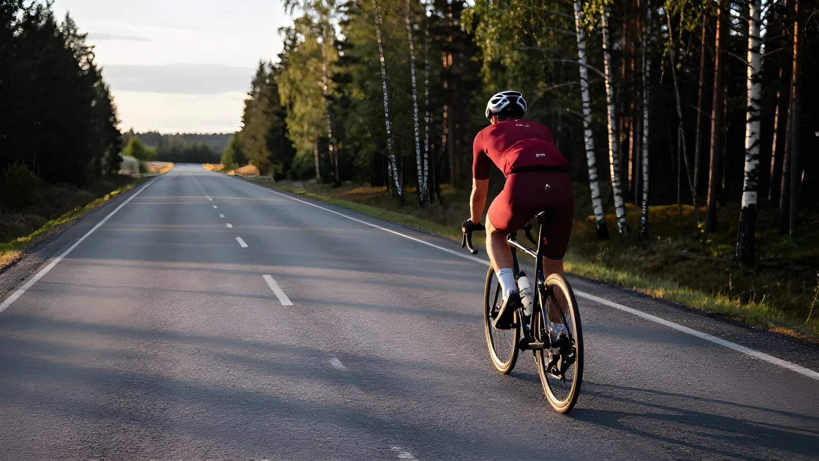 Ensam landsvägscyklist i röd tröja cyklar på en tom svensk landsväg i gyllene kvällsljus med björkar och tallskog längs vägkanten.