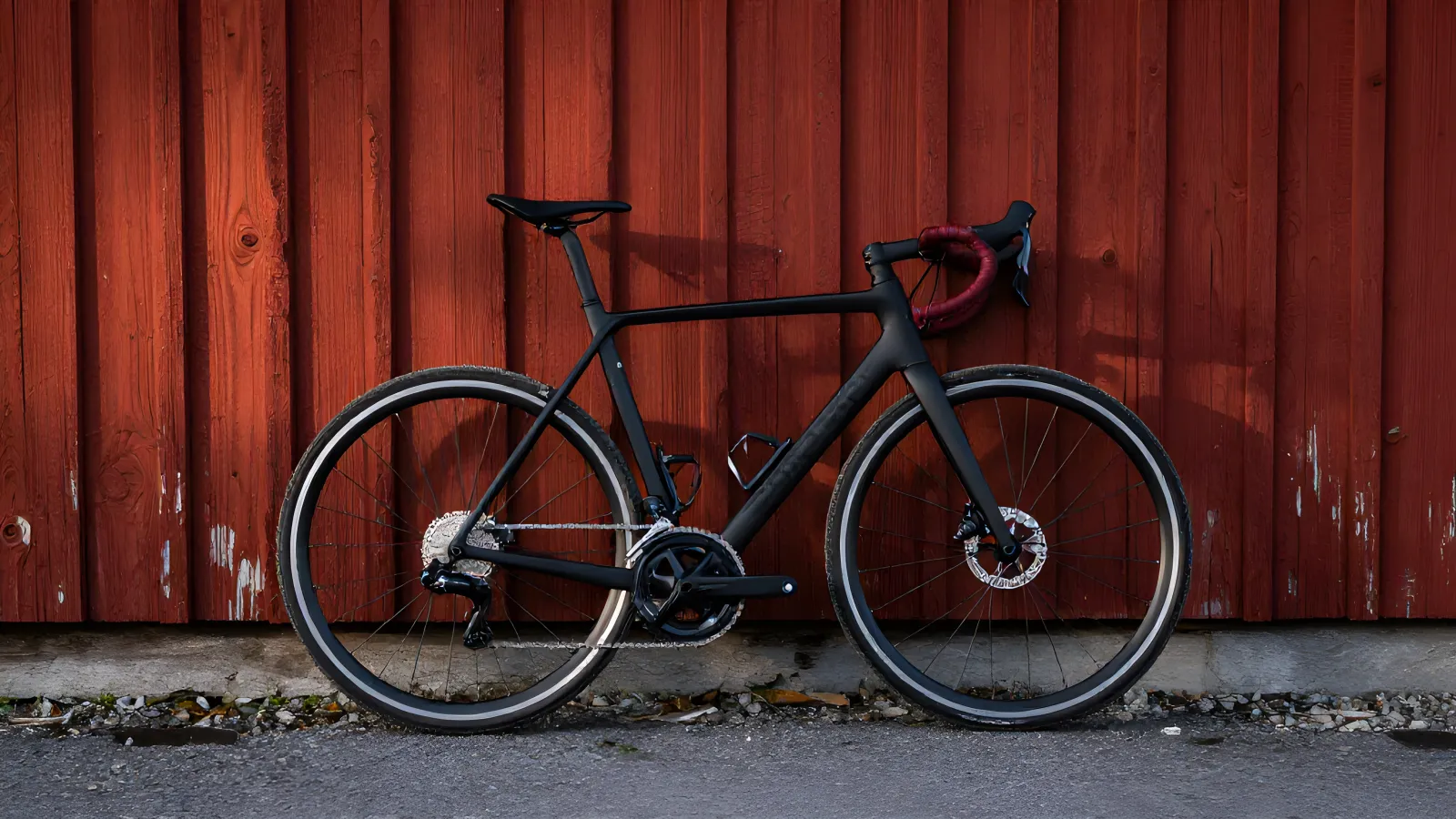 Racercykel med smala däck och skivbroms i sidoprofil, lutad mot en falurödfärgad trävägg i varmt kvällsljus.